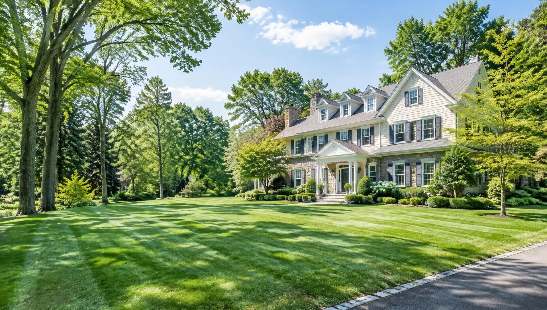 Cream shingle-and-stone colonial home with a thick, freshly striped front lawn and tall mature trees in Chatham, NJ