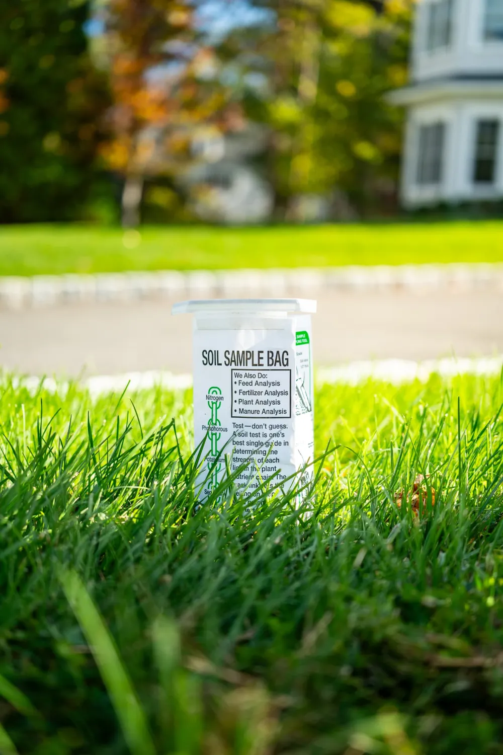 Laboratory soil sample bag standing in thick green grass on a front lawn with a white colonial home and autumn trees behind it in Northern New Jersey