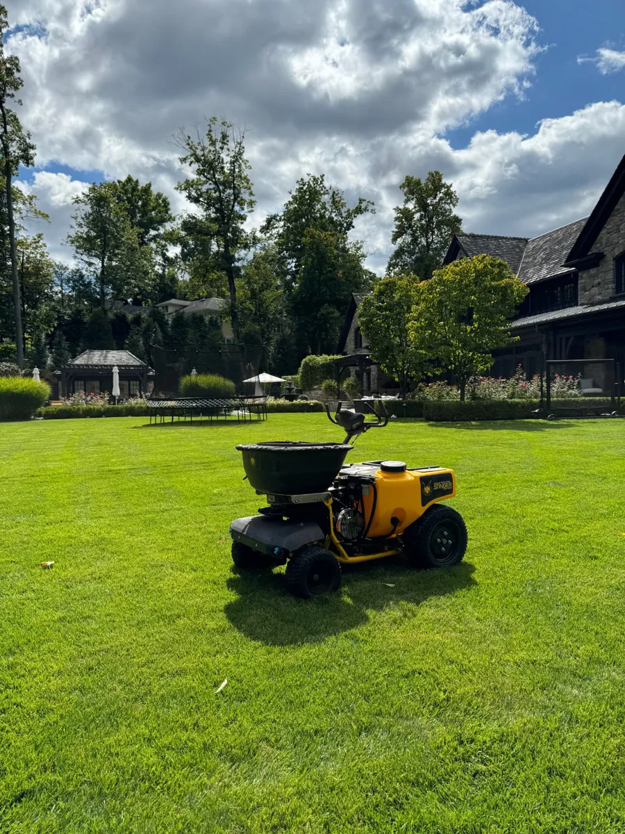 GreenStripe Stinger stand-on spreader-sprayer parked on a dense, bright green backyard lawn behind a stone estate on Beechwood Road in Summit, NJ, treated on the GreenStripe Superintendent Lawn Program