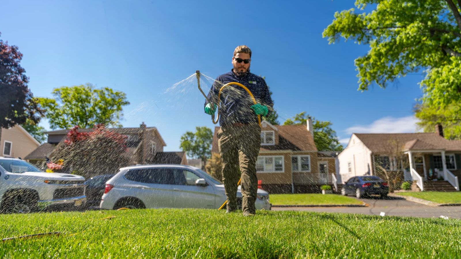 GreenStripe lawn technician spraying a liquid lawn treatment across a thick green front lawn on a residential street in Kenilworth, NJ