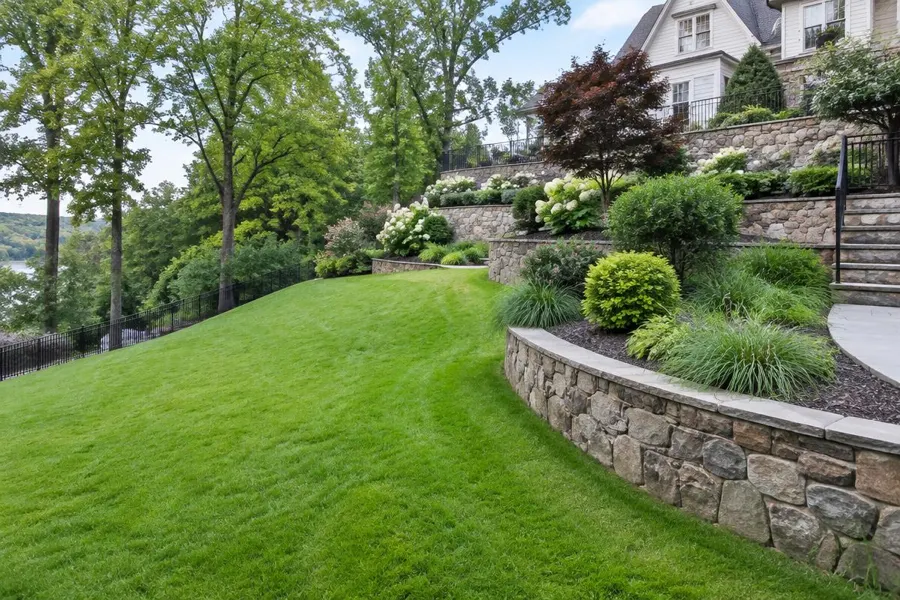 Sloping lawn beside curved stone retaining walls and terraced planting beds on a hillside property in Short Hills, NJ