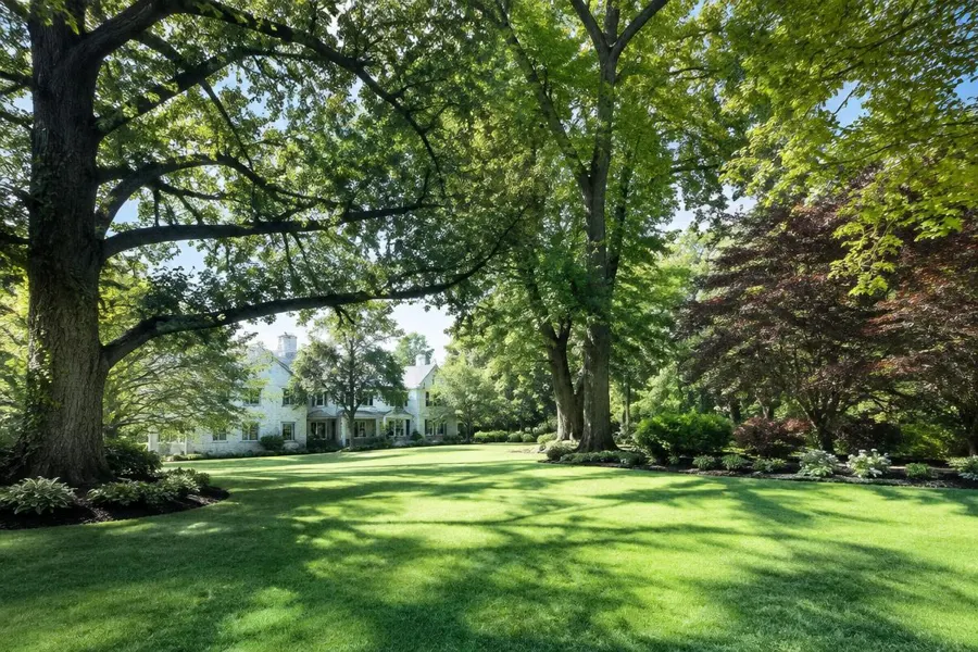 Expansive lawn in dappled shade beneath large mature trees with ornamental beds and a white home in the distance in Short Hills, NJ