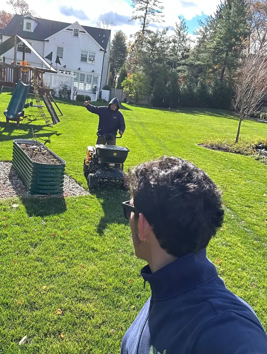 GreenStripe technician applying granular fertilizer with a stand-on spreader across a thick, striped backyard lawn behind a white colonial home on Prospect Street in Summit, NJ, on the GreenStripe Superintendent Lawn Program