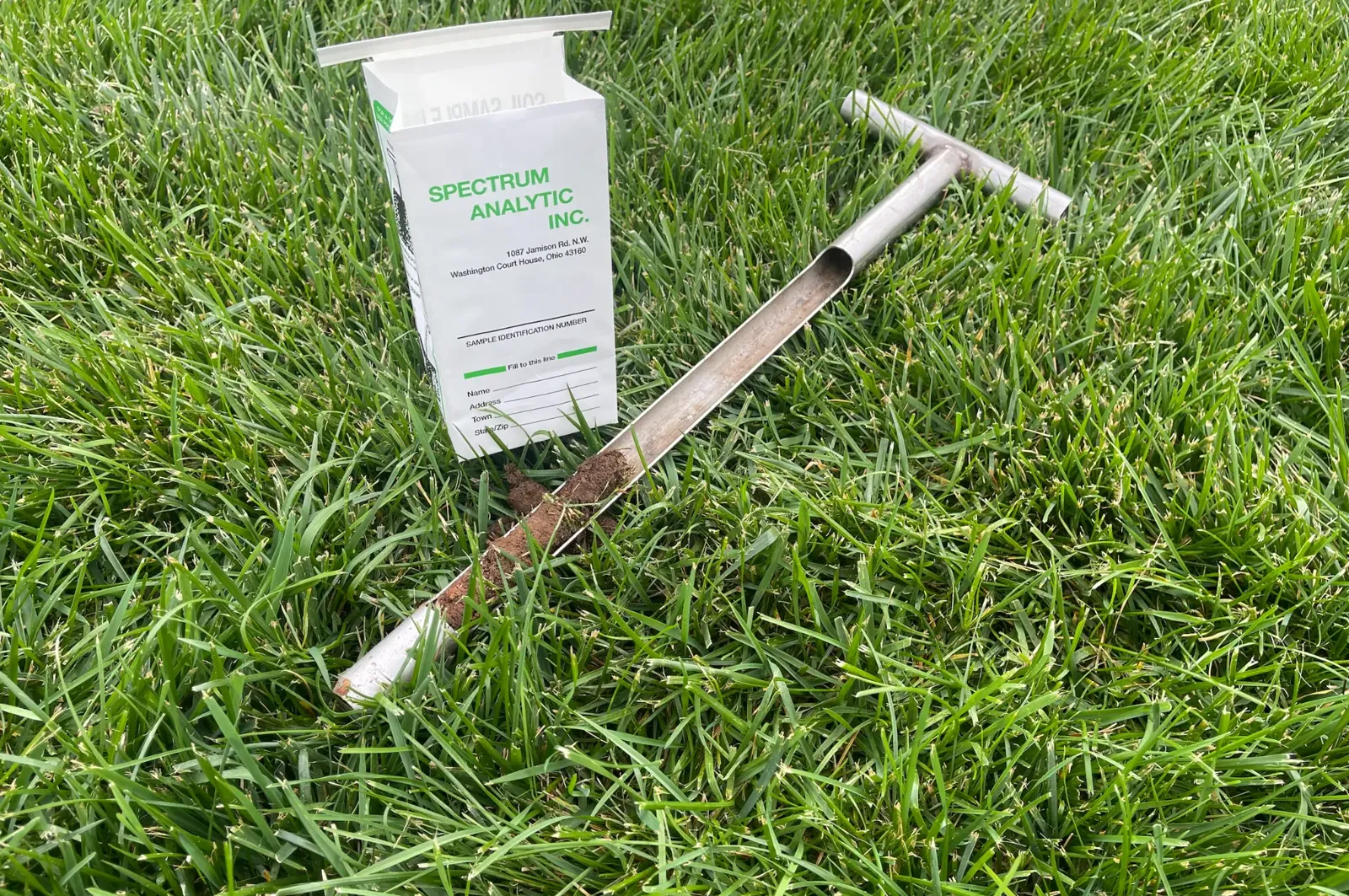 Soil probe with a fresh core sample next to a soil test bag on a lawn